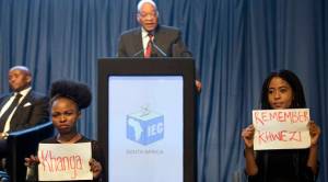 Protesters hold up papers protesting against President Jacob Zuma, at podium as he delivers a speech at the announcement of the results of the municipal elections in Pretoria, South Africa, Saturday, Aug. 6, 2016. The protest refers to Zuma's acquittal for rape in 2006. (AP Photo/Herman Verwey)